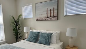 A bedroom with a bed, two wall-mounted lamps, a framed picture of surfers, and a potted plant. - ViBe District Retreat #2 (Virginia Beach)