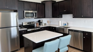 A modern kitchen with dark cabinets, stainless steel appliances, a central island with chairs, and a white tiled backsplash. - ViBe District Retreat #2 (Virginia Beach)