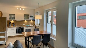 A compact kitchen with modern appliances, a dining area with a wooden table and chairs, and a large window with blinds. - Strandwiese - Ankerplatz - Ankerplatz - Strandnah... Wellenrauschen Inklusive! (Dahme)