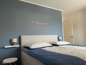 A bedroom with a bed, bedside lamps, a small table, and a wall with a decorative sign.
