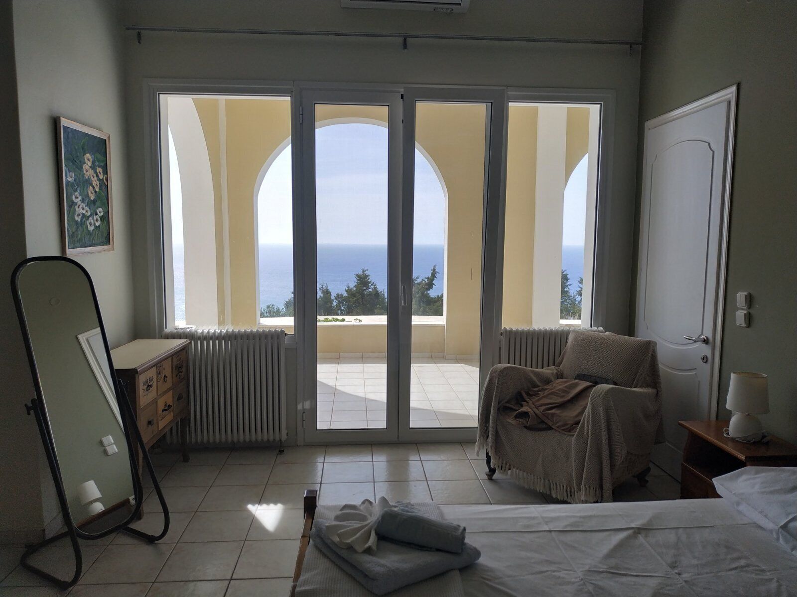 A bedroom with a bed, a chair, a dresser, a mirror, and a view of the sea through large glass doors.