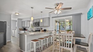 Een moderne keuken met grijze kastjes, een centraal eiland met krukken, een plafondventilator en een eethoek met tafel en stoelen.