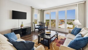 Ein modernes Wohnzimmer mit Flachbildfernseher, Essbereich mit Stühlen, einer Sofagarnitur und Blick auf eine entfernte Stadtlandschaft durch große Fenster.