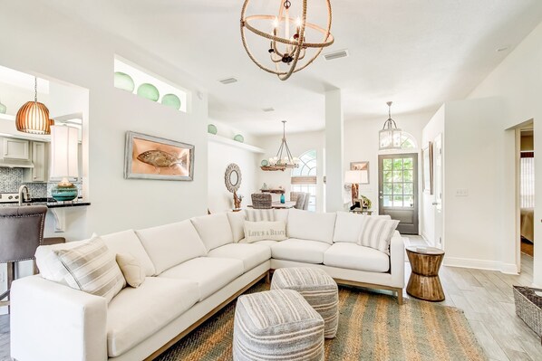 A spacious living room with a large L-shaped sectional sofa, a chandelier, and a dining area with a kitchen in the background.