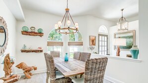 Un comedor con una mesa de madera y sillas de mimbre, una araña y objetos decorativos en estantes.