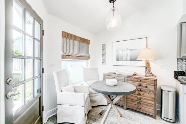 A room with a white door, a round table with a white bowl, a wooden dresser, and a lamp.