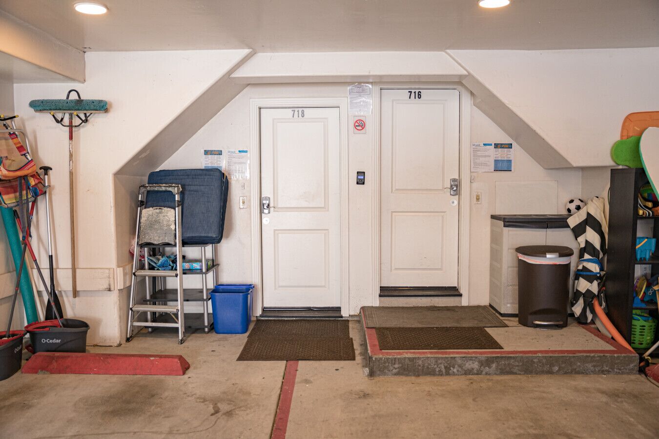 Un petit garage fermé, doté de deux portes blanches, d’une poubelle bleue, d’une échelle en métal et de divers outils de nettoyage.