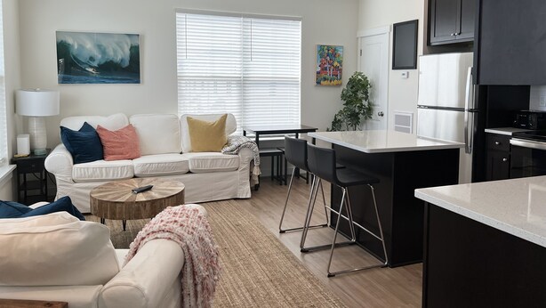 Una sala de estar moderna con un sofá blanco, una mesa de centro y una cocina con refrigerador, microondas y taburetes de bar.