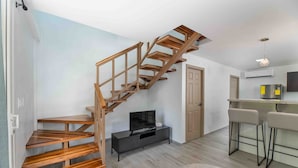 A modern living space with a wooden staircase, a flat-screen TV, a kitchen area with bar stools, and a door.