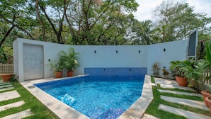 A rectangular swimming pool with blue mosaic tiles, surrounded by a concrete deck and potted plants.