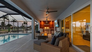 A covered outdoor area with a pool, wicker furniture, a ceiling fan, and a kitchen area with a TV. - Sauna, Paddle Boards, Gulf Access, Heated Pool - Villa Sun Kissed - Roelens Vacations (Cape Coral)