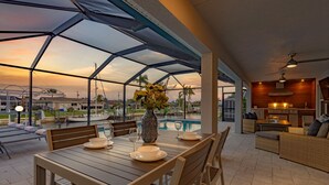 A covered outdoor dining area with a table set for two, a vase with flowers, and a view of the pool and sunset. - Sauna, Paddle Boards, Gulf Access, Heated Pool - Villa Sun Kissed - Roelens Vacations (Cape Coral)