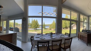 Un comedor con una mesa grande dispuesta para cuatro personas, ventanas amplias que ofrecen vistas a un lago y árboles, y una lámpara colgante moderna.