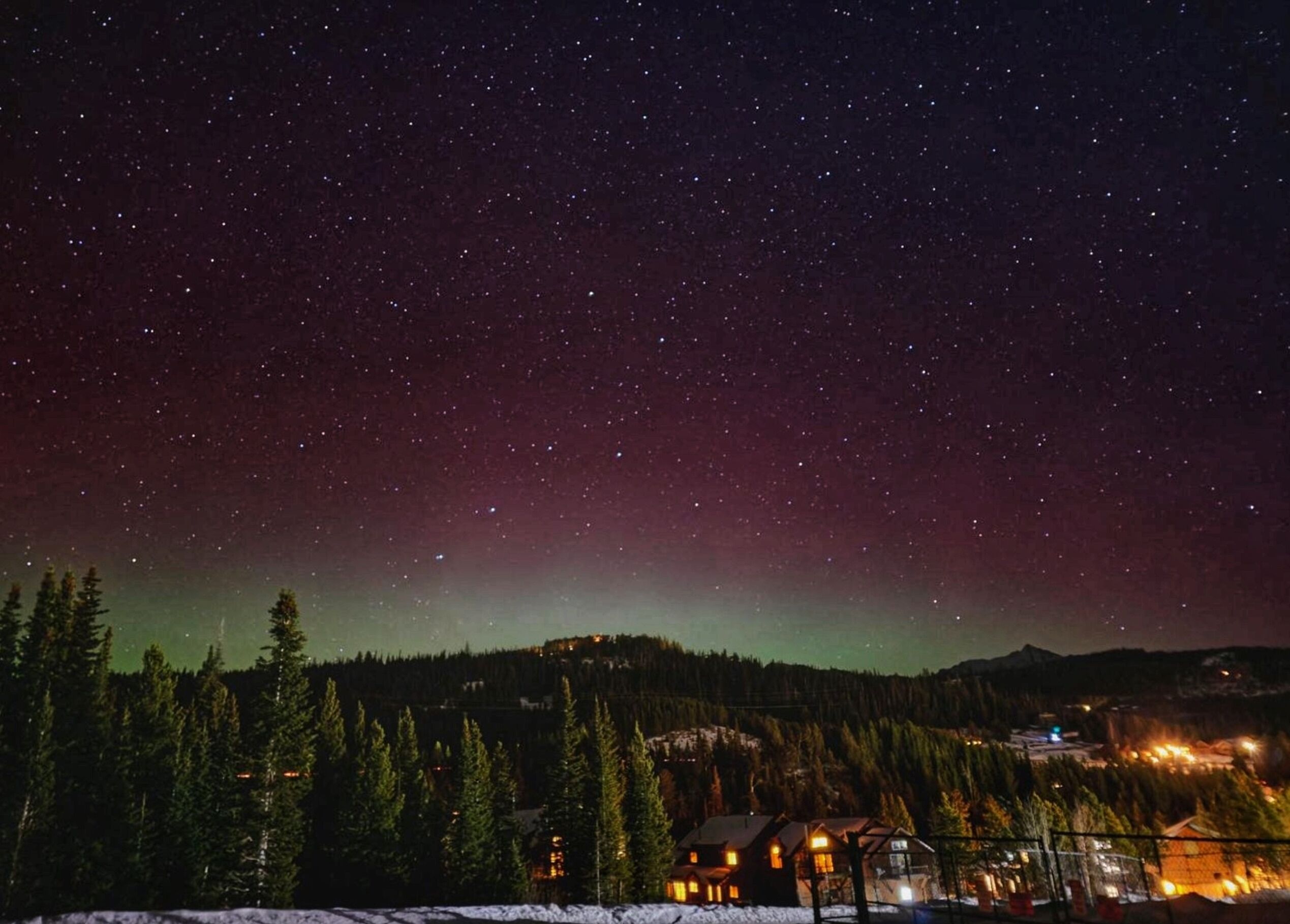A night sky with stars and a visible aurora. There is a house with lights on, surrounded by trees and a snowy landscape.