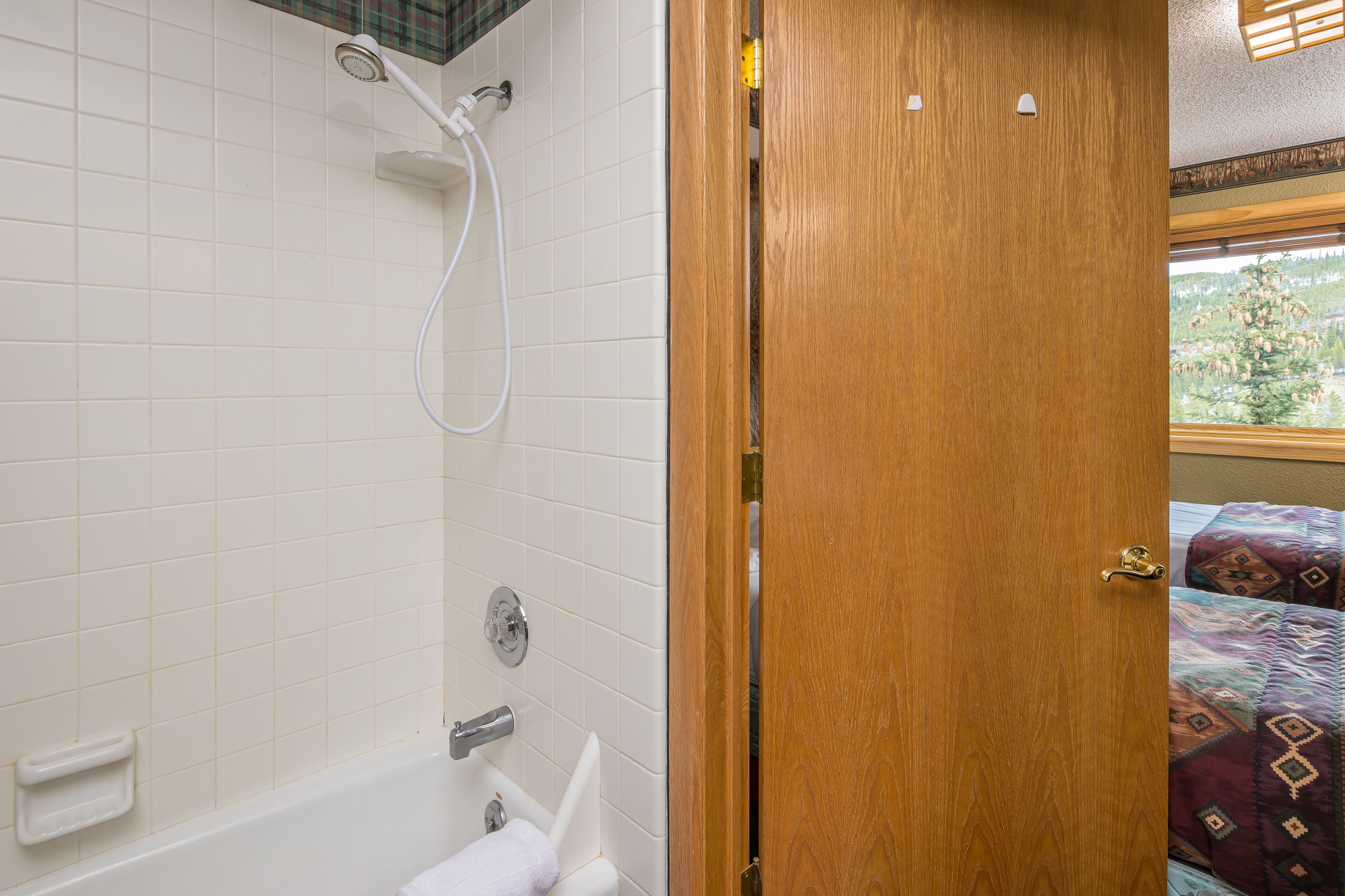 A bathroom with white tiles, a shower, a bathtub, and a wooden door leading to a room with a window.