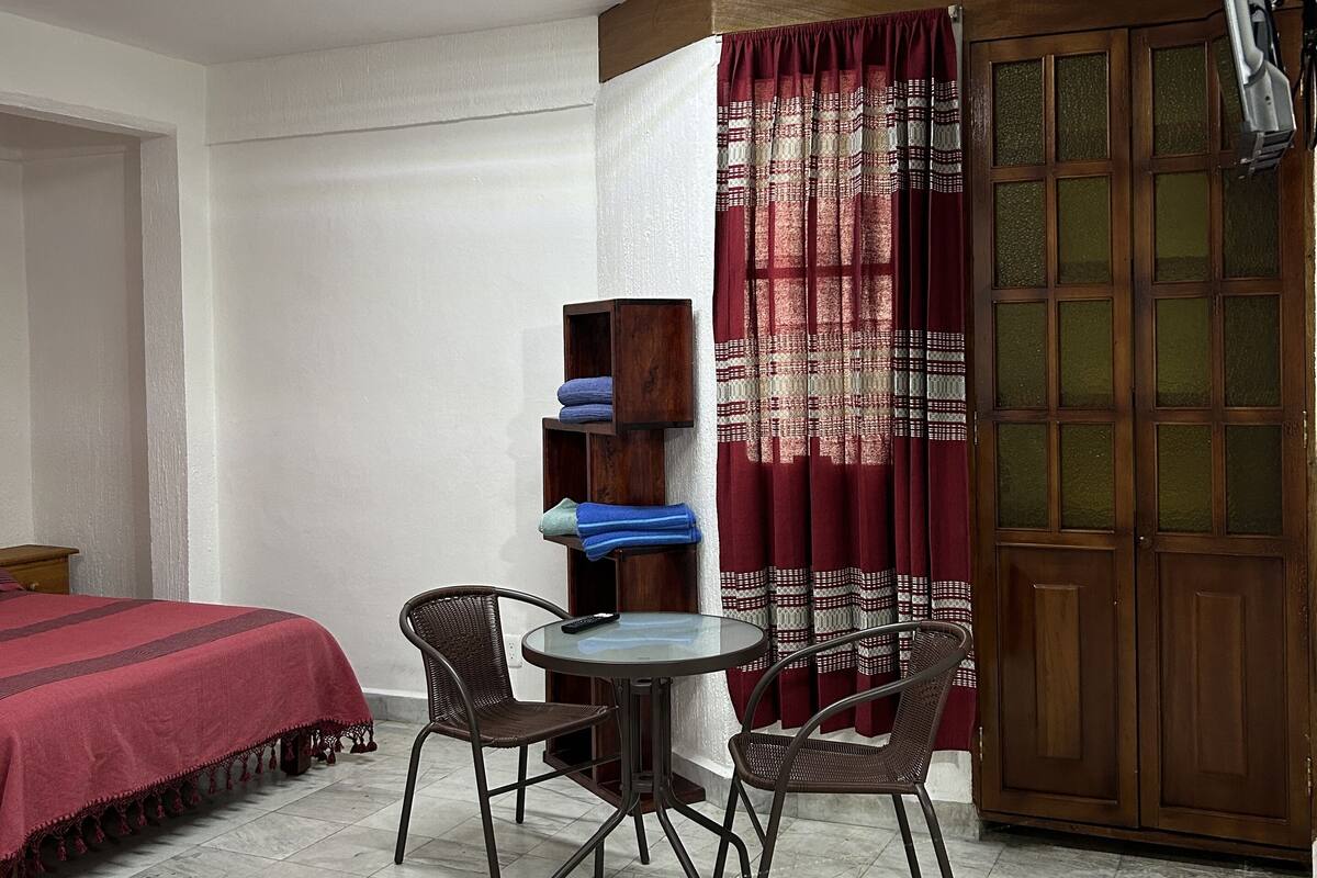 A small, well-lit room with a bed, a table with chairs, a wooden wardrobe, and a window with curtains.