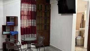 Una habitación pequeña y bien iluminada con una mesa redonda y dos sillas, un armario de madera con toallas dobladas, una cortina roja estampada y un televisor montado en la pared.