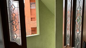 Un pasillo con puerta de madera que incluye un ventanal de vitral, que da a una escalera y una vista de un edificio con fachada verde.