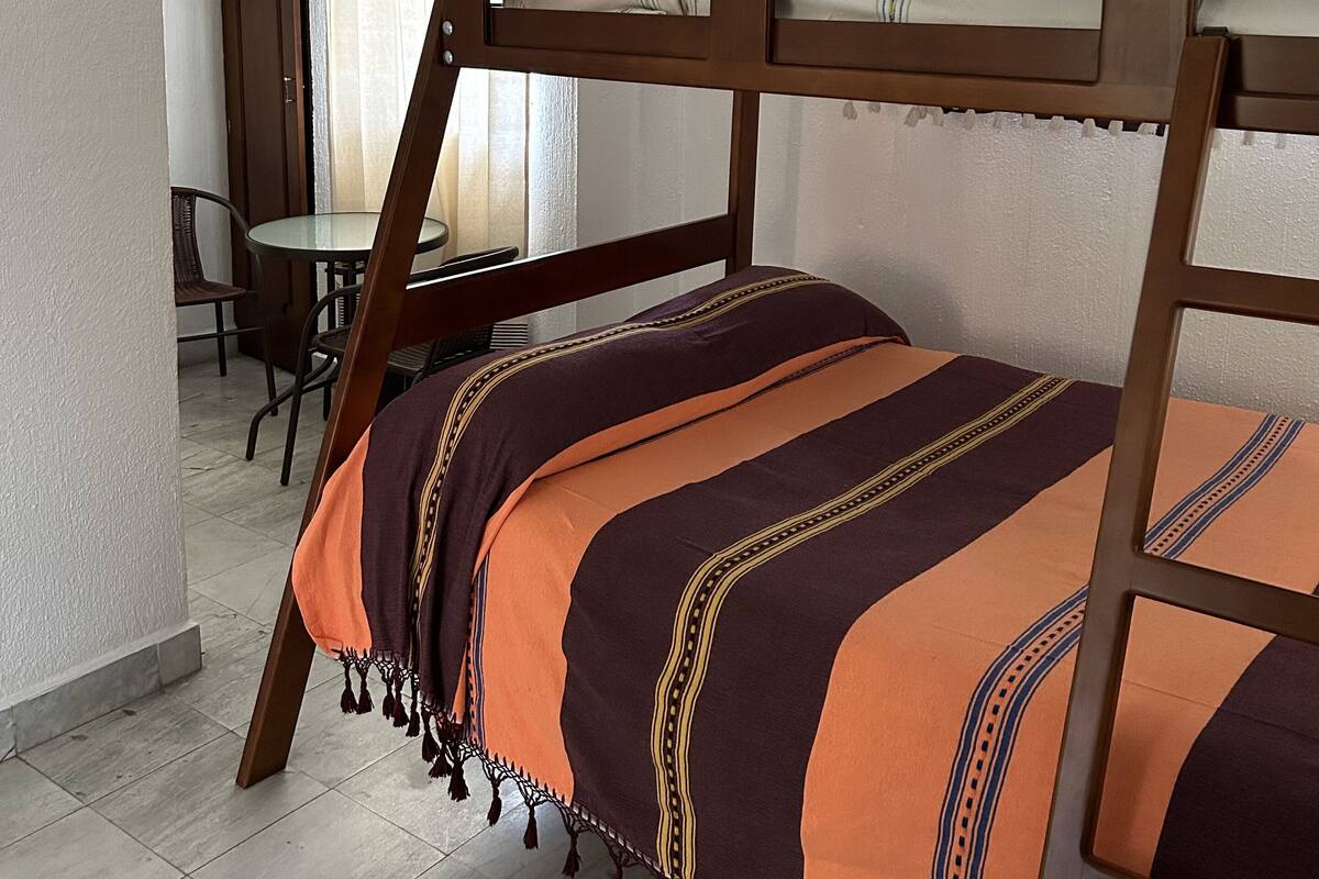 A single bed with a wooden frame and a striped blanket, a small table, and a chair in a room with white walls and a tiled floor.