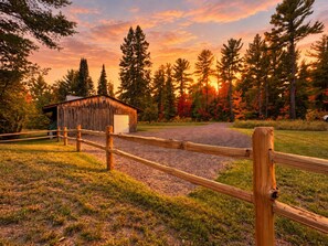 A rustic wooden barn with a gravel driveway leading to it, surrounded by a wooden fence and a variety of trees.