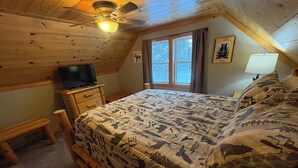 Ein Schlafzimmer mit Holzbalkendecke, einem Bett mit gemusterten Betttextilien, einem Fernseher auf einer Holzkommode und einem Fenster mit Vorhängen.