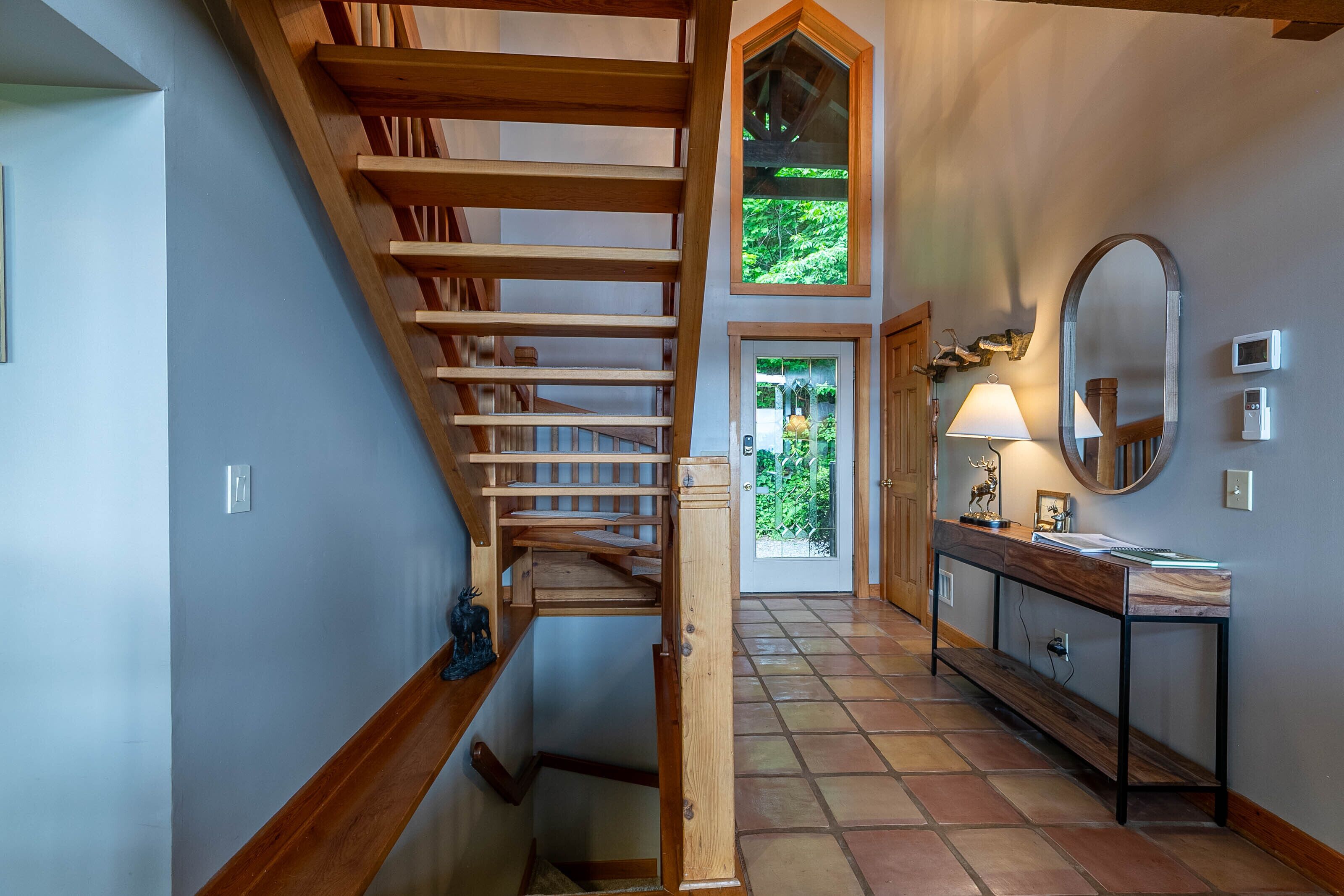 A staircase leading to a second floor, a console table with a mirror, and a door with a view of greenery outside.