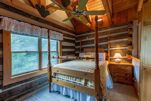 A log cabin bedroom with a four-poster bed, a wooden nightstand with a lamp, and a window with a view of trees.