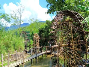 Un puente de madera rústico que conecta dos grandes norias en un paisaje verde y exuberante, con montañas al fondo.