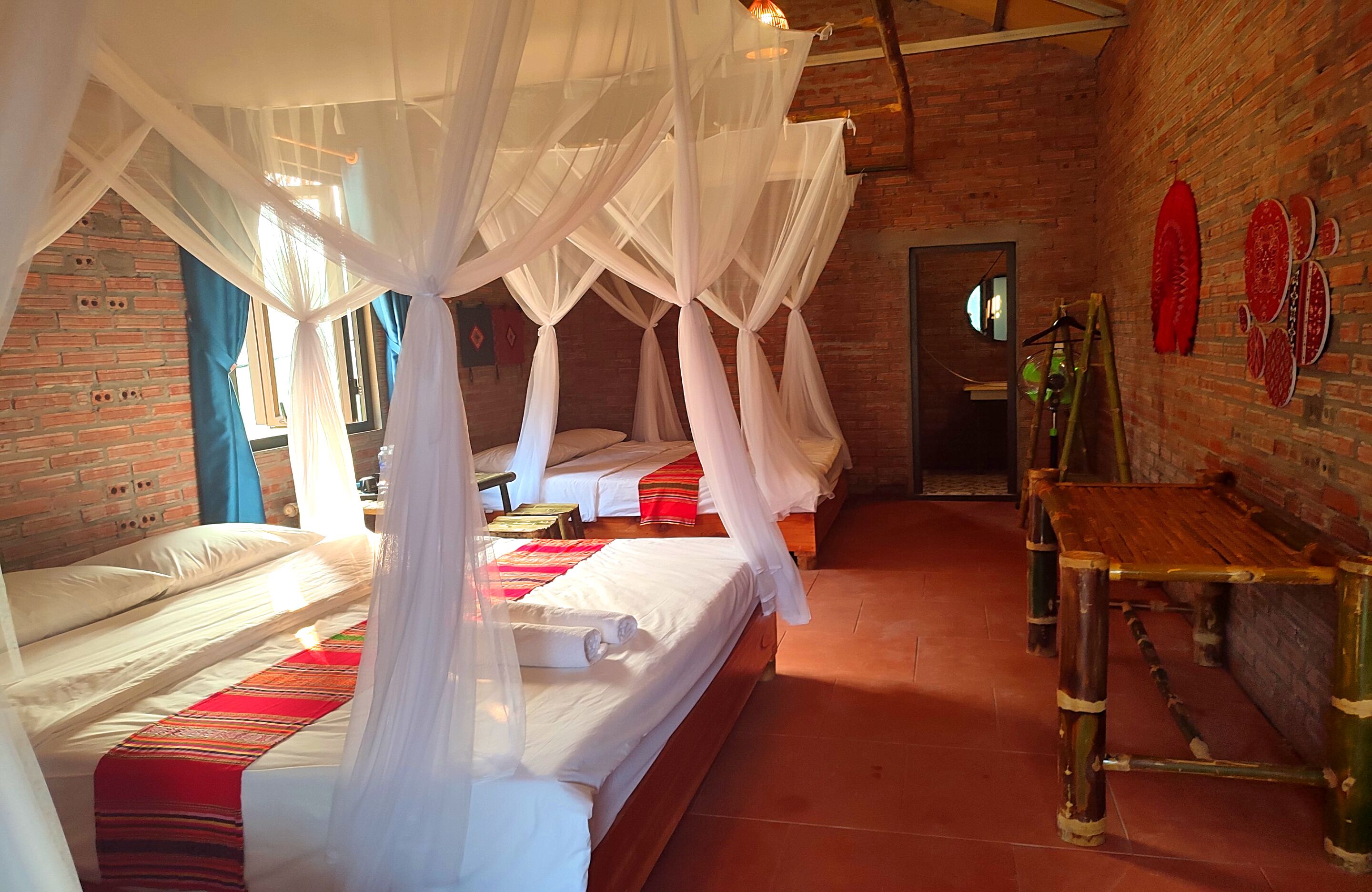 A room with four beds, each with white linens and a red and white striped blanket. There is a wooden bench and a small table with a green bottle. The room has a brick wall and a large window with blue curtains.