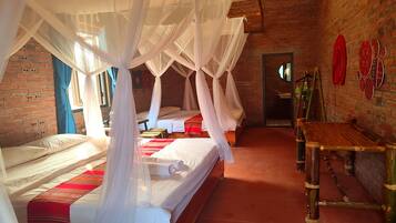 A room with four beds, each with white linens and a red and white striped blanket. There is a wooden bench and a small table with a green bottle. The room has a brick wall and a large window with blue curtains.