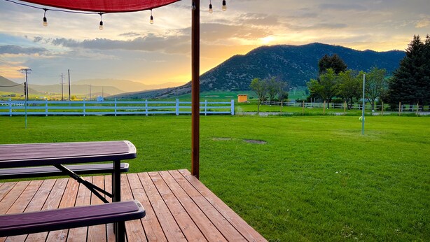A wooden picnic table under a red canopy with string lights, set against a backdrop of a grassy field, white fence, and mountains. - Townside@LavaHotSprings (Lava Hot Springs)
