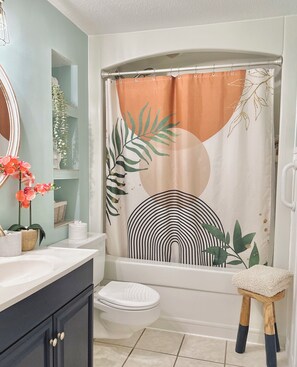 A bathroom with a white bathtub, a white toilet, a wooden stool, a mirror, and a shelf with decorative items.