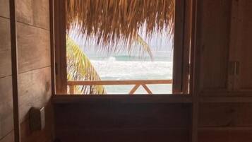 Een houten kamer met een bed, een raam met rieten dak en uitzicht op de oceaan.