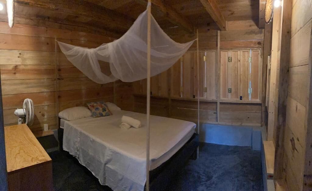 Un dormitorio de cabaña de madera con una cama, una mesita de noche, un ventilador y una mosquitera.