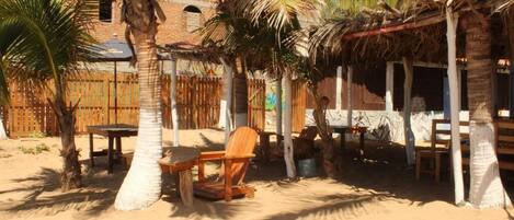 A beachside outdoor seating area with wooden tables and chairs, surrounded by palm trees and a thatched-roof structure.