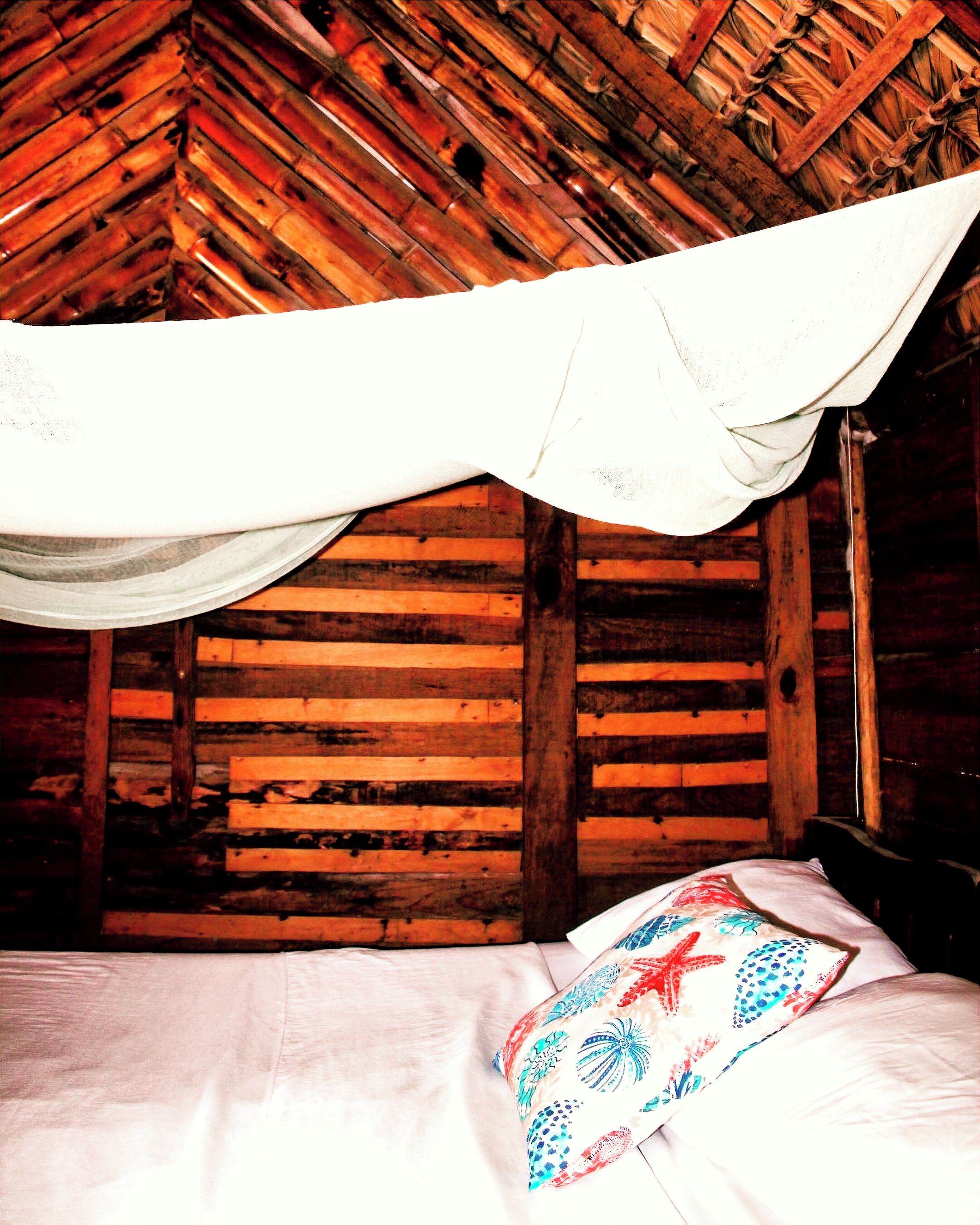 A room with a wooden ceiling and walls, a bed with a white sheet and a colorful pillow, and a white canopy over the bed.