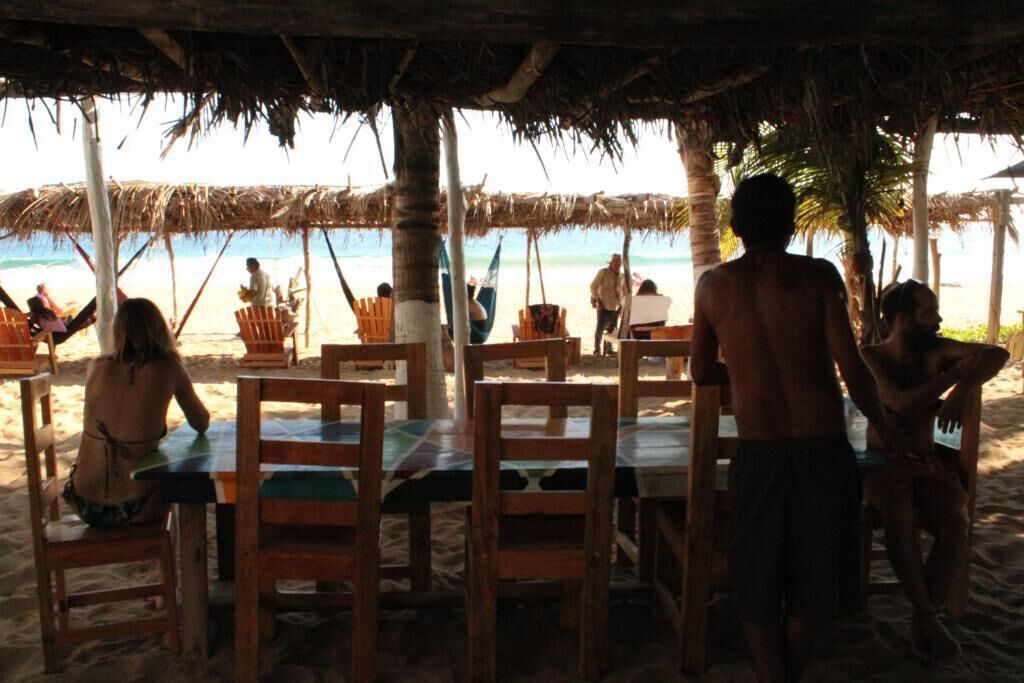 A beachside restaurant with wooden tables and chairs, a thatched roof, and a view of the ocean.