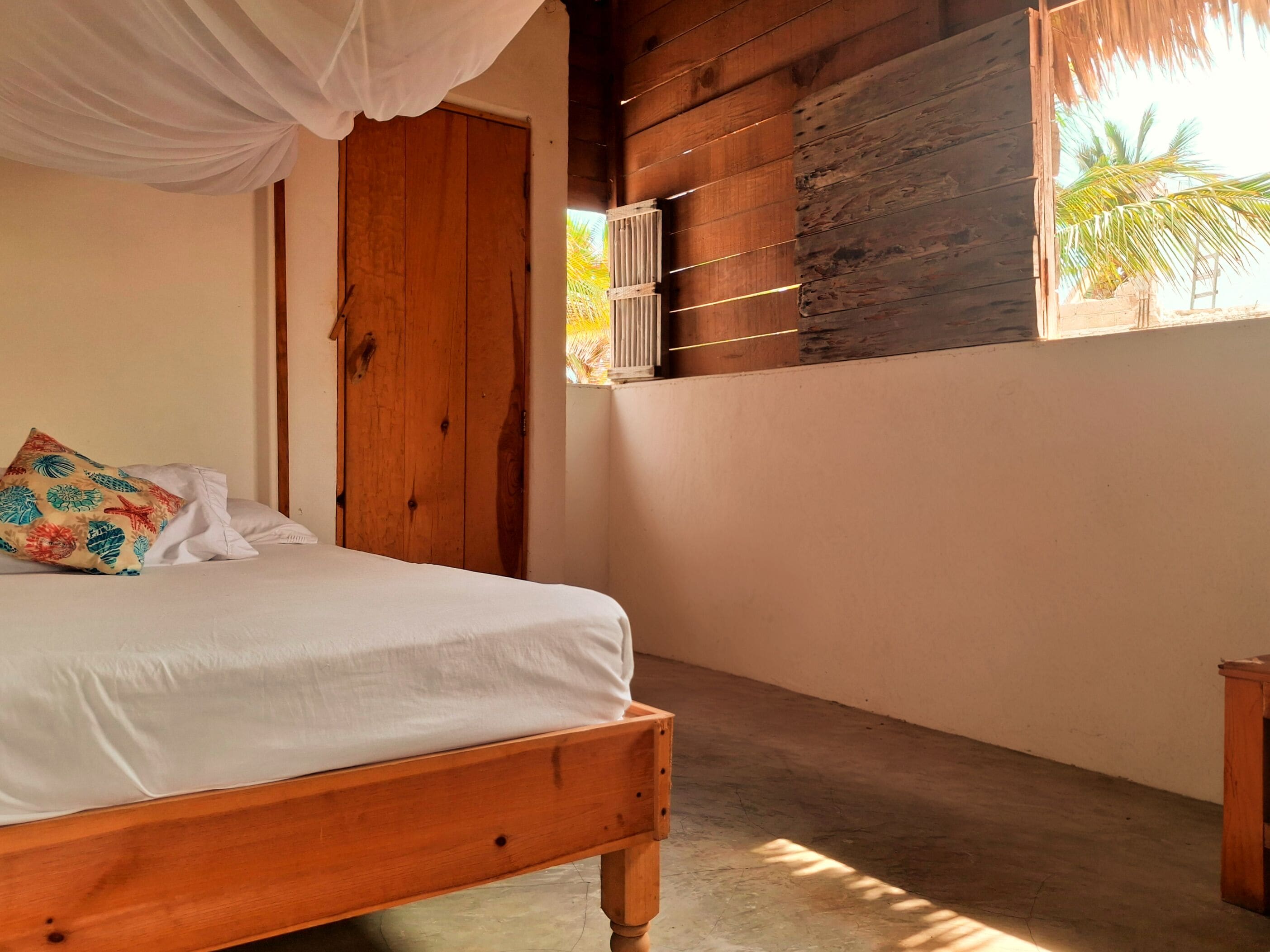 A bedroom with a wooden bed, a white canopy, a wooden door, and a view of palm trees through a window.