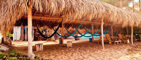 Una cabaña en la playa con techo de paja, pilares de madera y una hamaca.