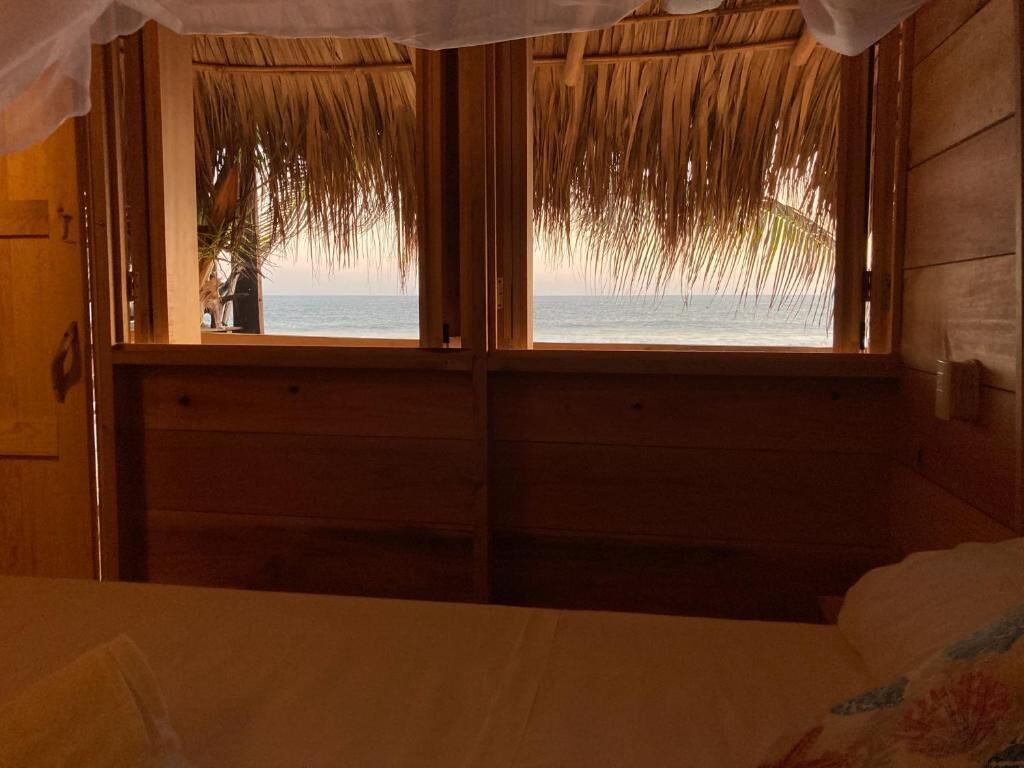 Una habitación con cama, paredes de madera, una ventana con vista al mar y un techo de paja.