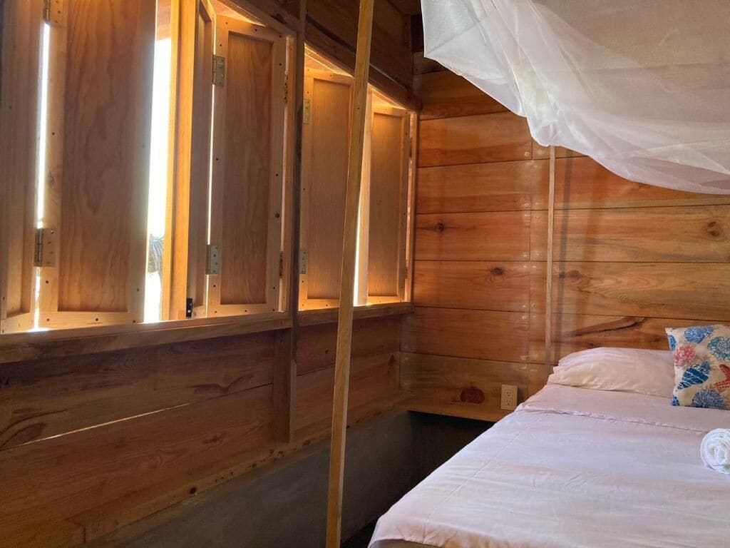 A wooden cabin room with a bed, a window with a curtain, and a bedside table.