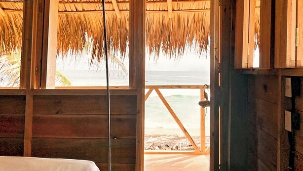 Una habitación de madera con una cama, una ventana con vista a la playa y un techo de paja.