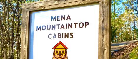 A wooden sign with the text "MENA MOUNTAINTOP CABINS" and a graphic of a cabin, welcoming visitors.