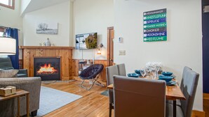 A cozy living room with a fireplace, a dining area with a table set for four, and a mounted sign with various brand names.