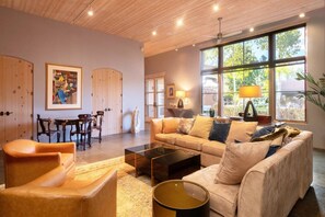 A modern living room with a sofa, coffee table, and chairs, a dining area with a table and chairs, and large windows with natural light.