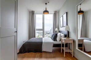 Et moderne soveværelse med et stort vindue, en seng med sort og hvid dyne, et natbord med en lampe og udsigt over byen.