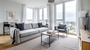 Un salon moderne avec un canapé gris, une table basse et un téléviseur posé sur un meuble en bois. De grandes fenêtres offrent une vue sur la ville.