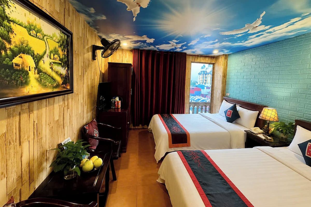 A hotel room with a mural, two beds, a desk, a chair, a plant, and a window with a view.