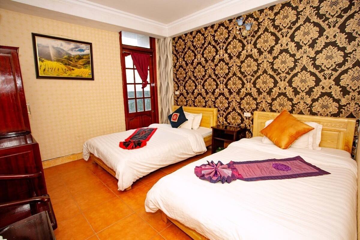 A hotel room with two beds, a wooden wardrobe, a window with curtains, and a patterned wallpaper.