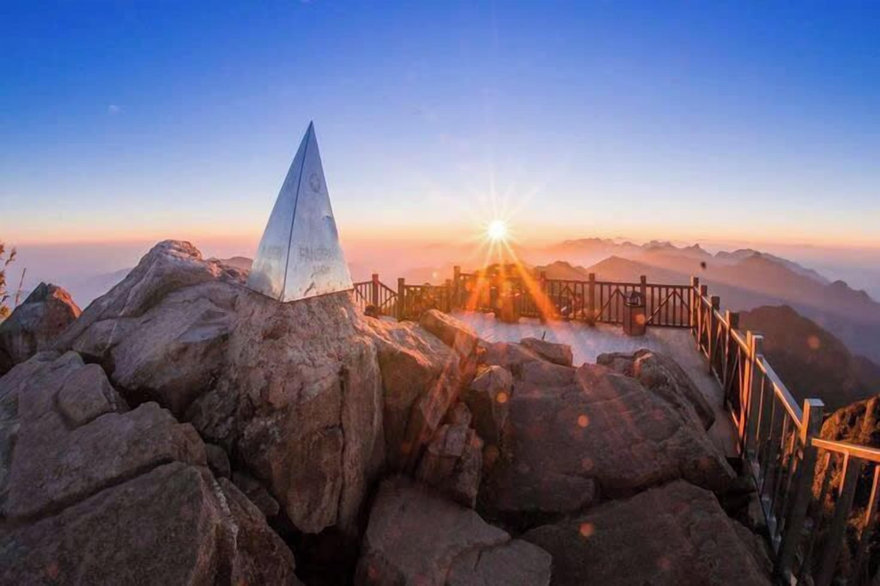 Un sommet de montagne surmonté d’un drapeau, une clôture en bois et un lever de soleil en arrière-plan.